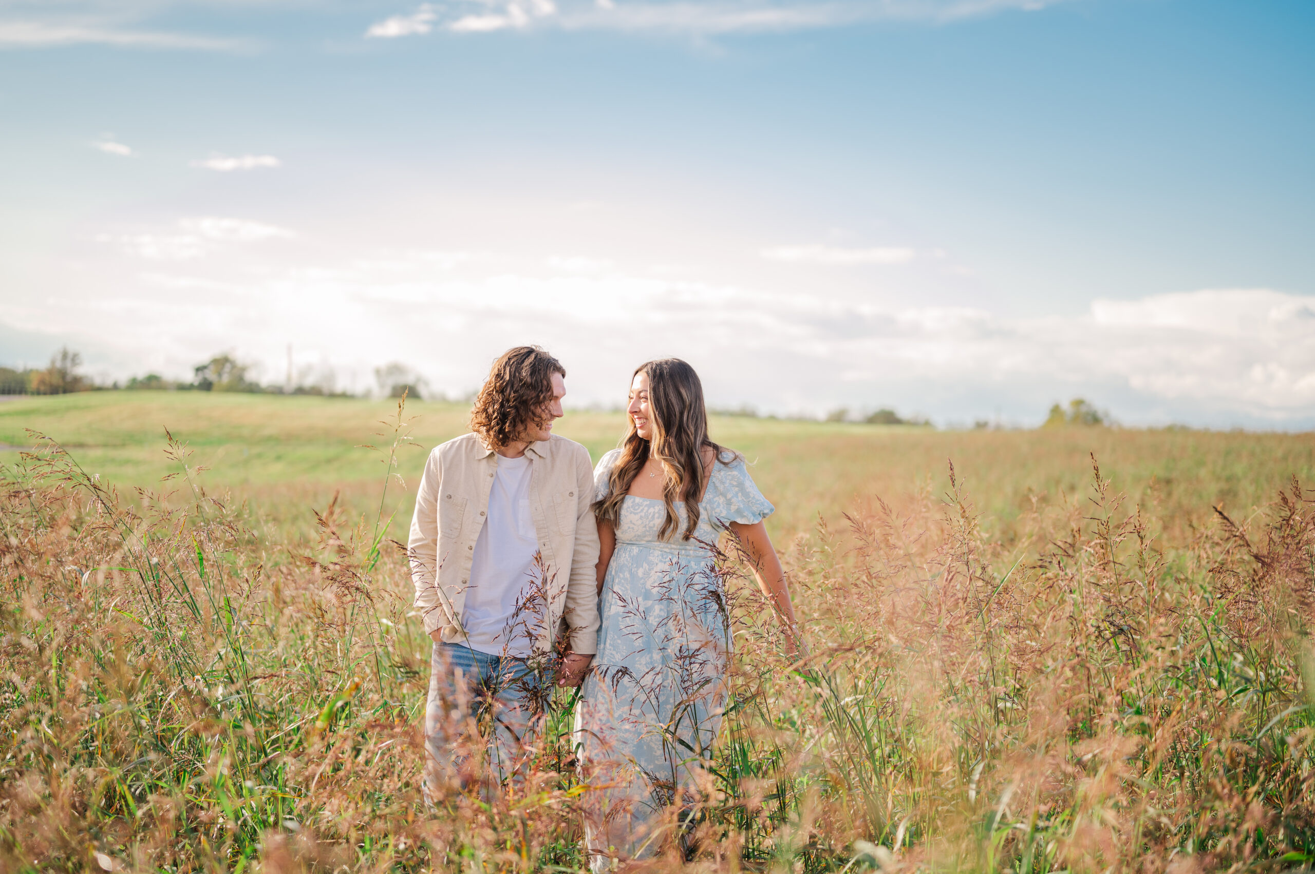 Couple walks through tall grass holding hands smiling at each other